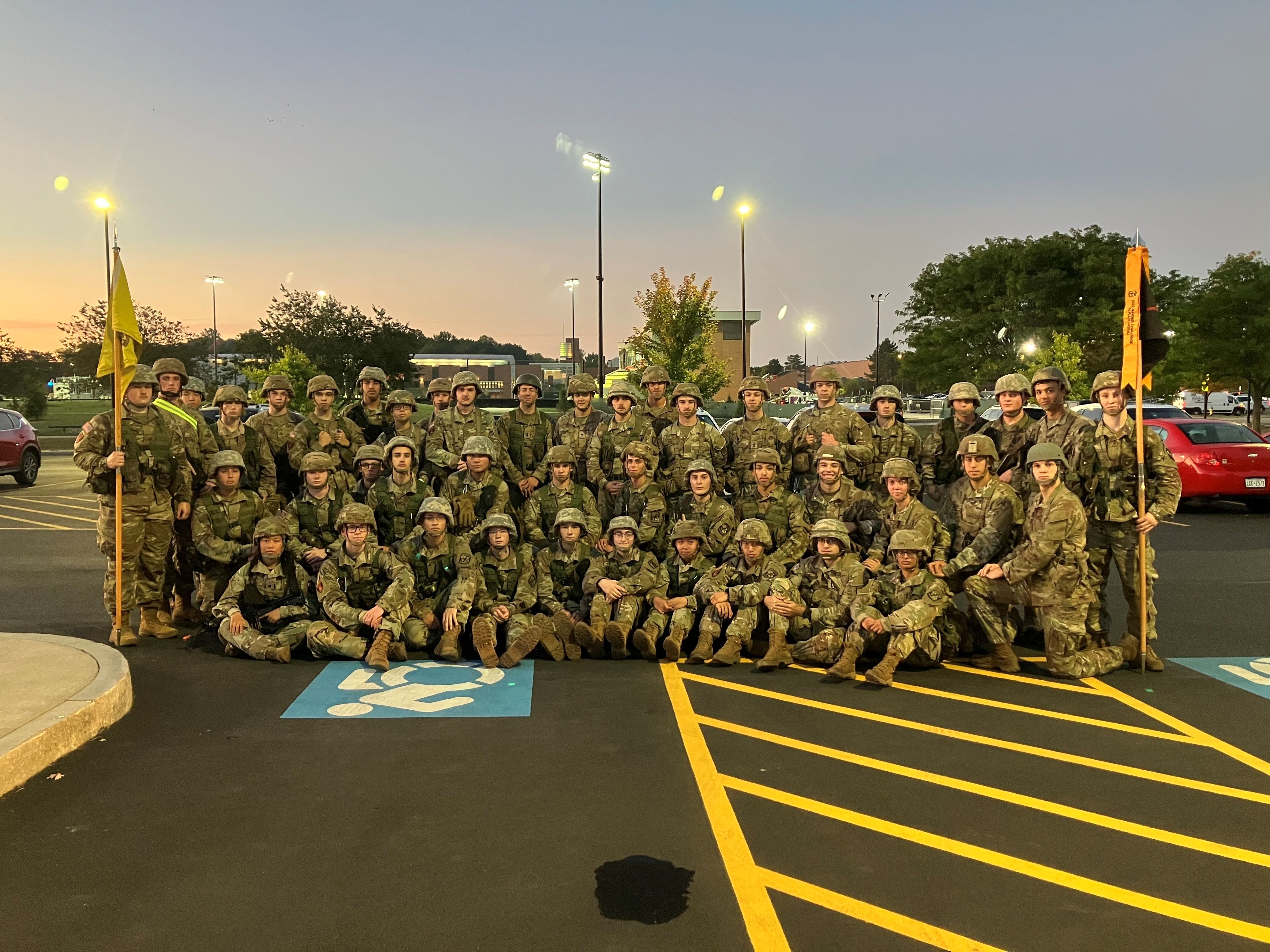 A large group of cadets wearing tactical vests and helmets and flanked by yellow guidon flags