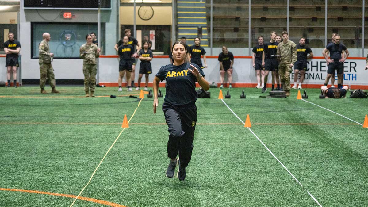 A female Army R O T C cadet sprints through an indoor training course with others watching.