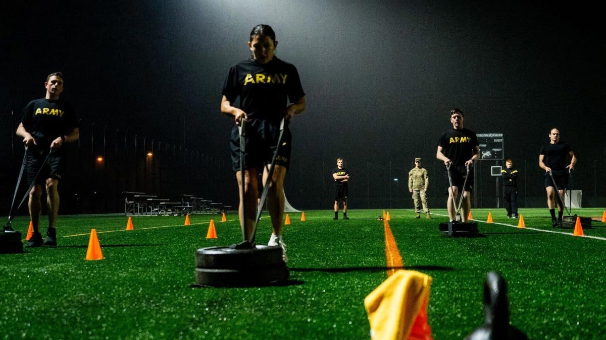 A female cadet is running backwards on a turf field, pulling a sled with 2 weight plates on it.
