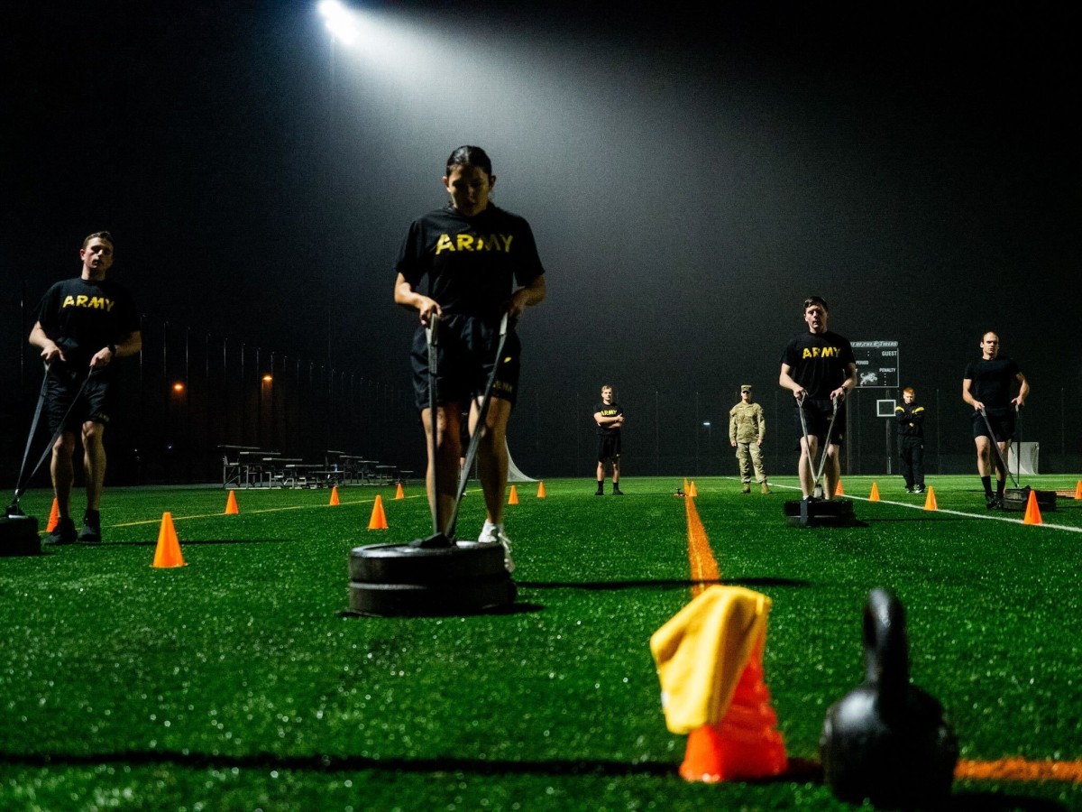 A female cadet is running backwards on a turf field, pulling a sled with 2 weight plates on it.