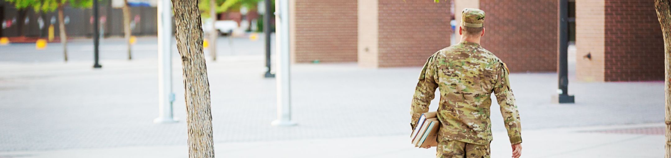 Man wearing an army uniform, walking on campus holding books.
