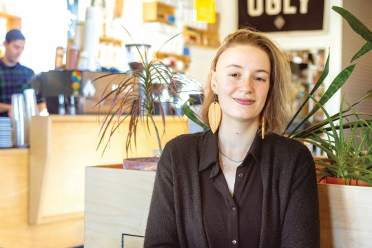 a college age student with mid-length blond hair and a black shirt sits in front of a plant in an office environment.