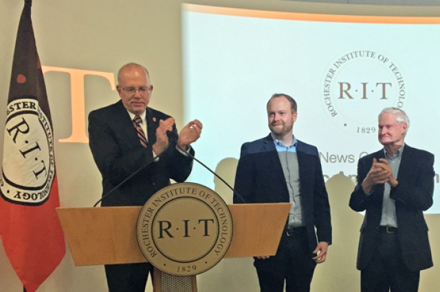 Person clapping standing behind a podium, accompanied by two other people.