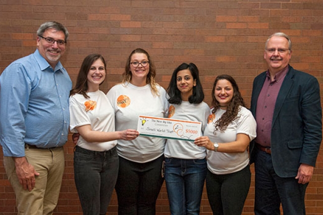 six people standing against a brick wall, with two holding a novelty bank check.
