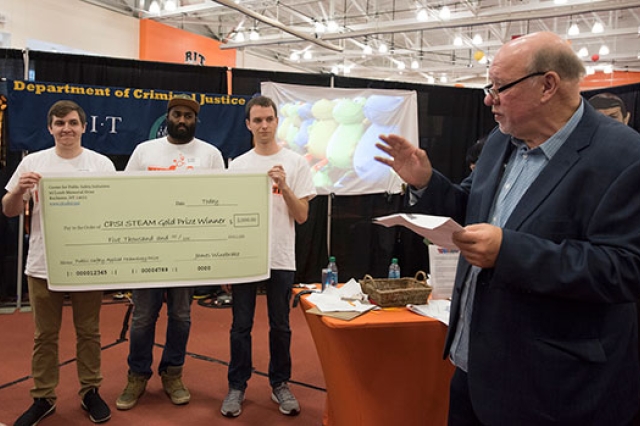Three Clockwyse team members hold up a large check while the CPSI director talks to a crowd about their first-prize accomplishment.
