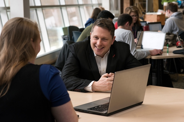 Michael Oshetski sits with a student in front of a laptop smiling.