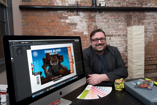 David Moffittt poses for a photo at his office desk. His computer is turned so the screen is facing the camera and the design for the beer label is pulled up on photoshop.