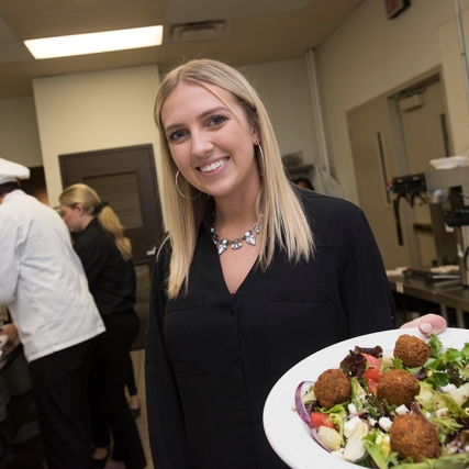 Student stands in restaurant kitchen holding plate of salad.