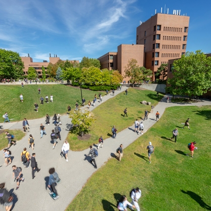 Students walking across walkway and lawn between brick buildings.