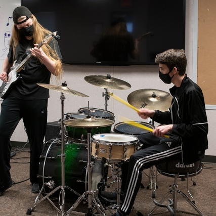 student playing the electric guitar next to student playing drums.