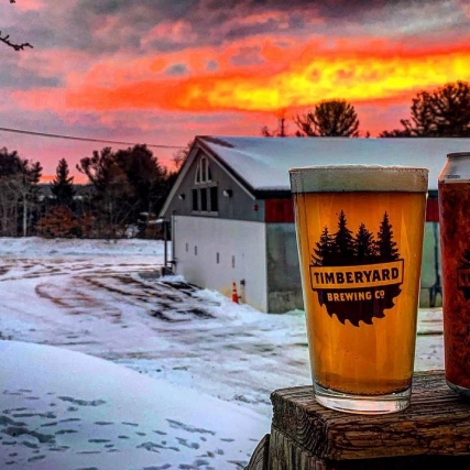 can and glass of beer in foreground of snowy scene with brewery in background.
