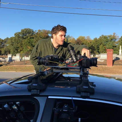 A person operates a camera rig on top of a car.