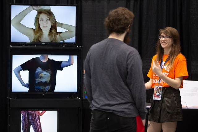 two students talking next to three stacked TV screens showing a person's head, torso, and legs.