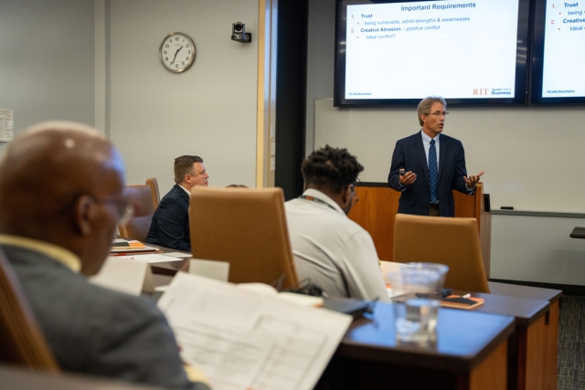 a man in a suit speaks to a room full of students with a projected display behind him.