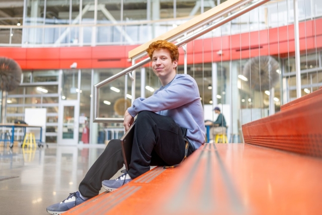 a young man with red hair and casual clothing sits on an orange staircase in a modern, brightly lit building.