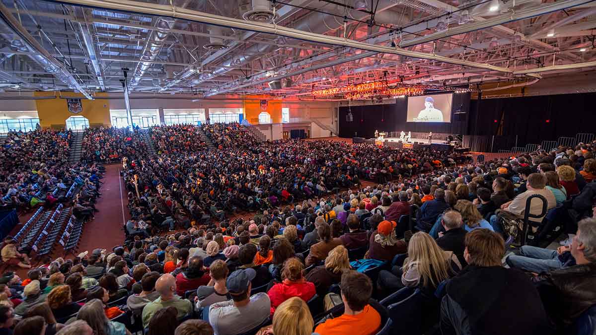A large indoor event in a packed field house with a speaker on stage and a big screen projecting the presentation.