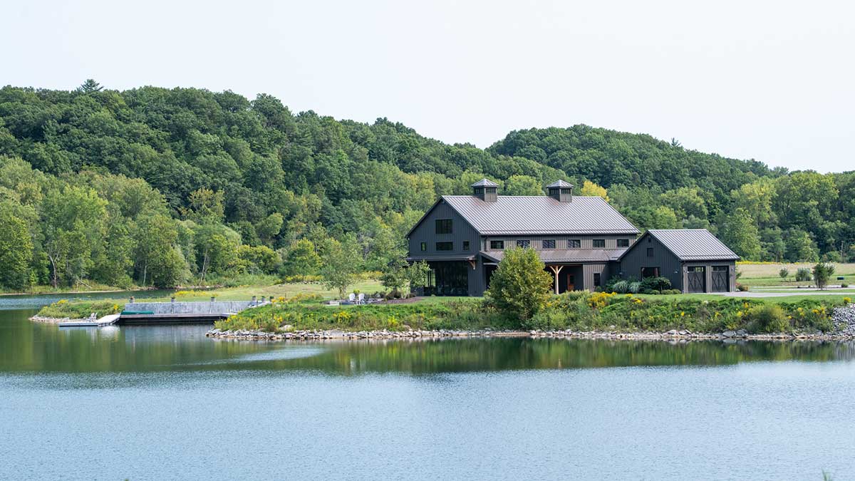 A modern barn-style building with a metal roof sits on a grassy shoreline next to a calm pond surrounded by green trees.