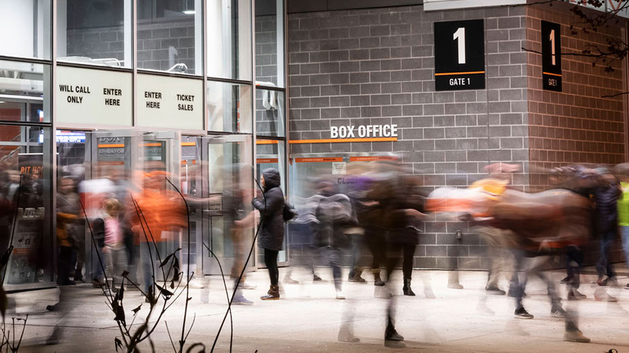 A blurred crowd of people enters through the glass doors at the box office of Gate 1 at night.