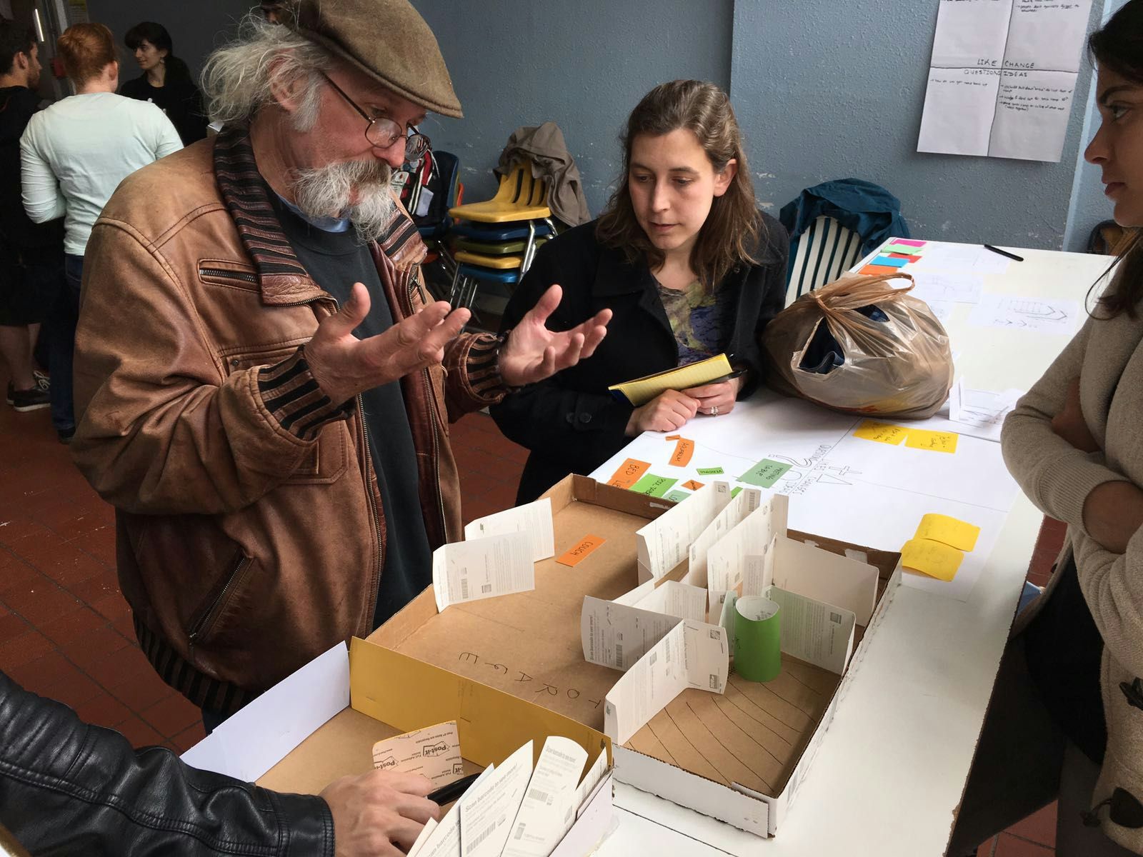 A group discusses a cardboard architectural model at a design workshop, with one man animatedly explaining while others listen.