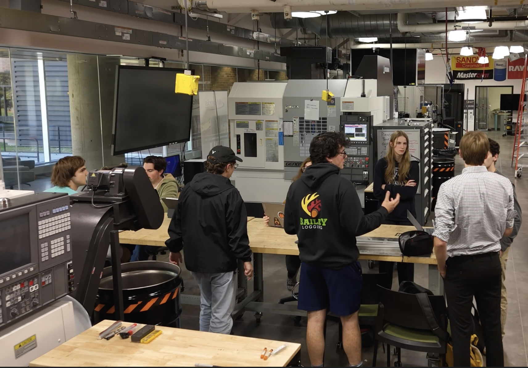 Students gather around a workbench in a modern machine shop, collaborating near CNC equipment and computer stations.