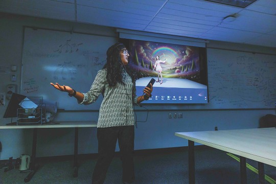 a female stands in a dimly lit classroom holding a phone in one hand. A screen showing a virtual reality game is in the background.