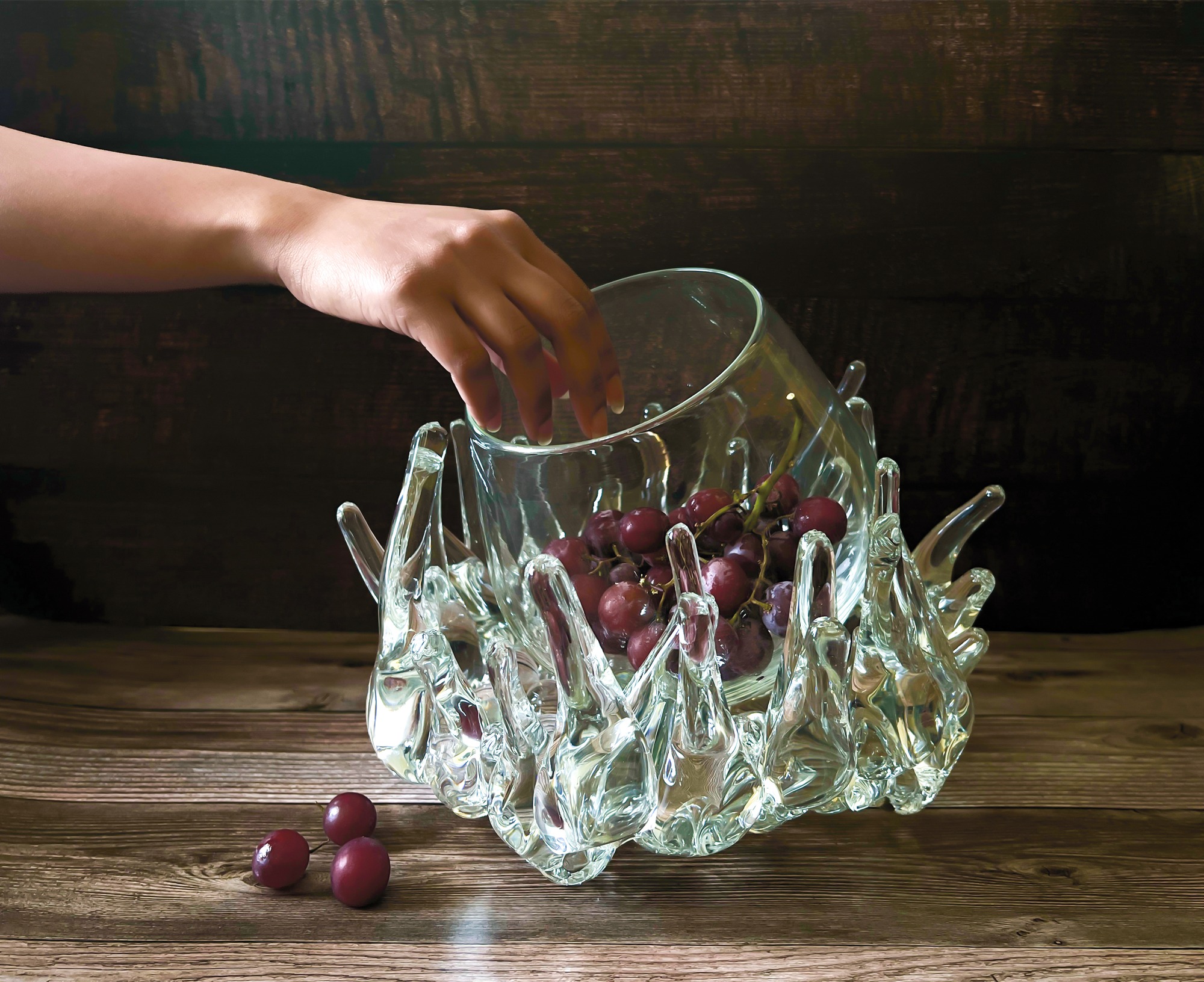 A hand reaches into a clear glass bowl supported by a sculptural glass base.