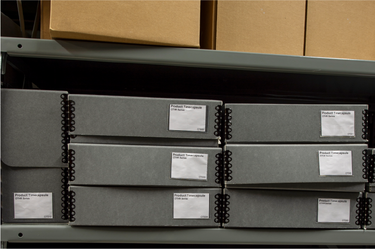 Shelf with neatly arranged gray binders labeled 'Product Timecapsule' alongside cardboard boxes.