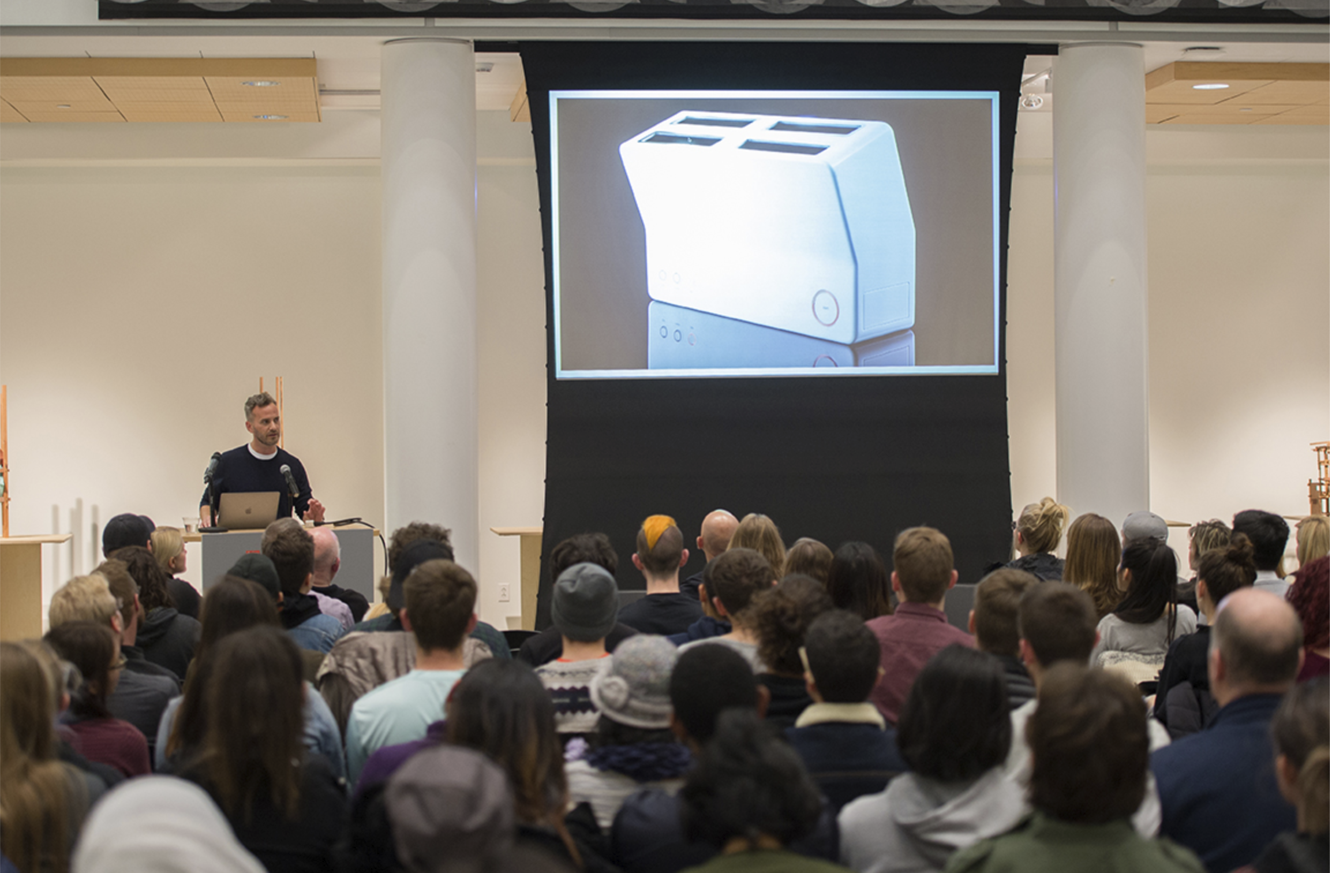A speaker presenting to an audience with a large projection screen displaying an image of a product behind him.