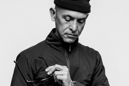 a black and white photo of a man holding glasses looking down at his phone. He wears a black shirt, a watch, and has a beanie on his head.