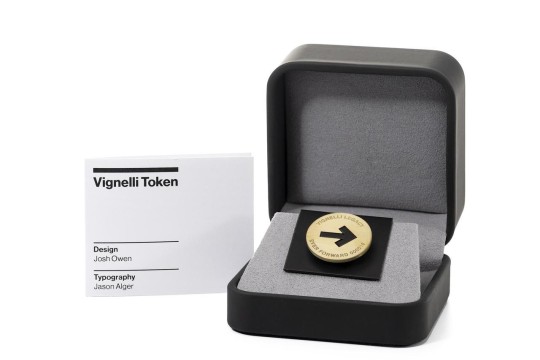 A brass Vignelli ‘Token of Inspiration’ displayed in an open gray presentation box, shown beside a card labeled ‘Vignelli Token’ with design credit to Josh Owen and typography credit to Jason Alger.