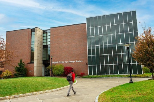 A student walks toward the Vignelli Center for Design Studies, a modern brick and glass building on a college campus, surrounded by green lawns and autumn trees.
