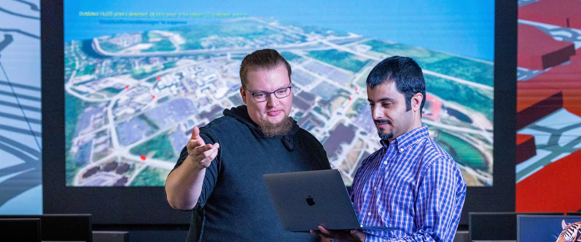 two researchers examining content on a laptop with a screen behind them showing a map.