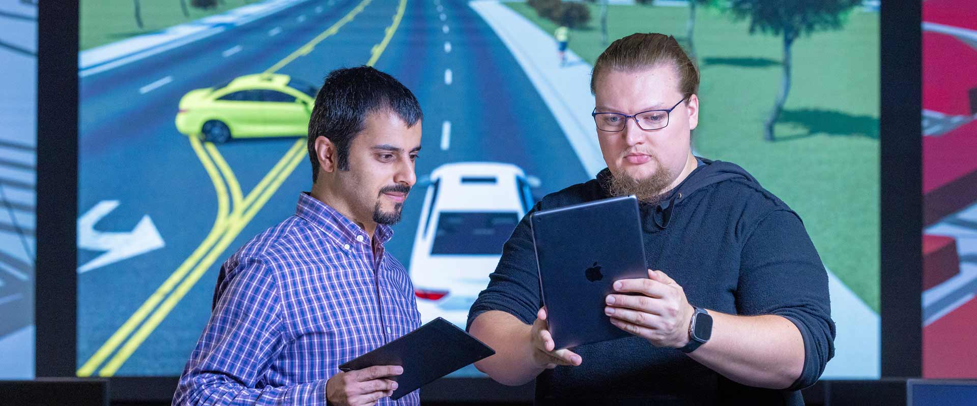 two researchers examining content on a laptop with a screen behind them showing an animation of cars on a roadway.