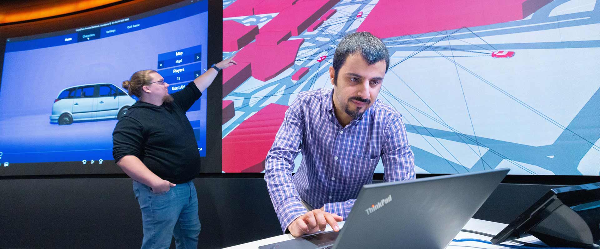 a researcher works on a laptop while another points to graphics on a screen in the background.