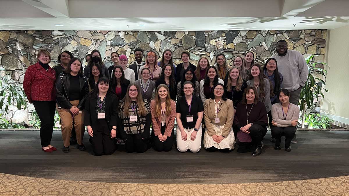 A professional group photo of students and mentors taken indoors against a stone wall.