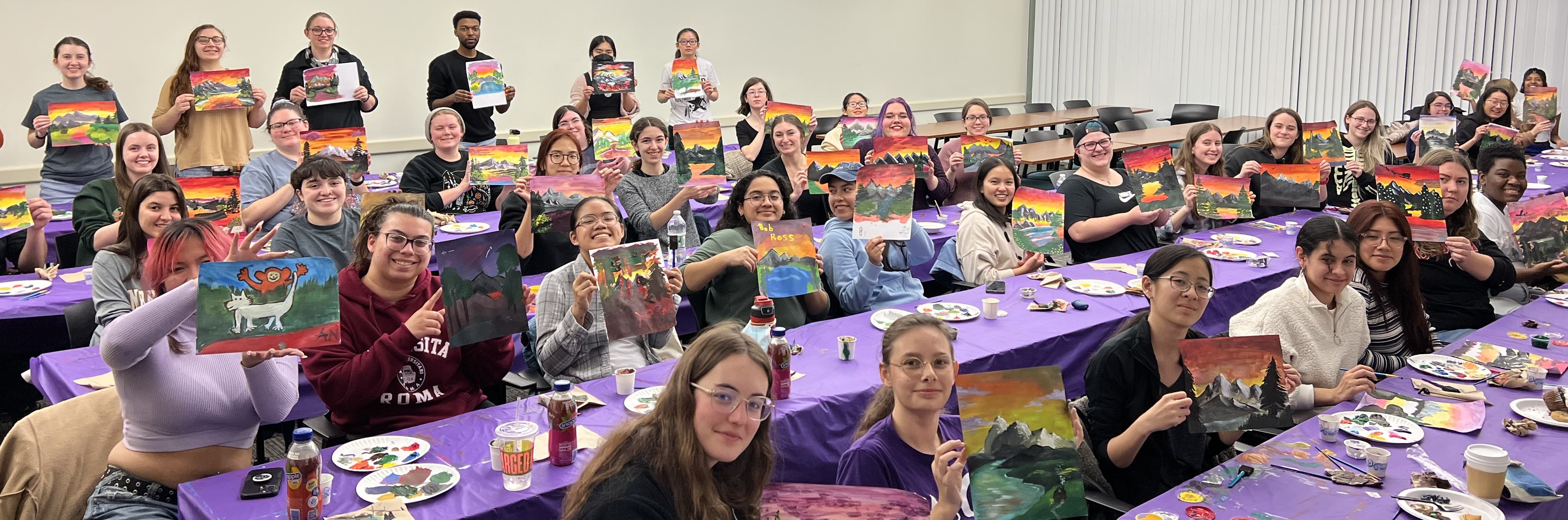 WiC Members holding up paintings at purple covered tables.