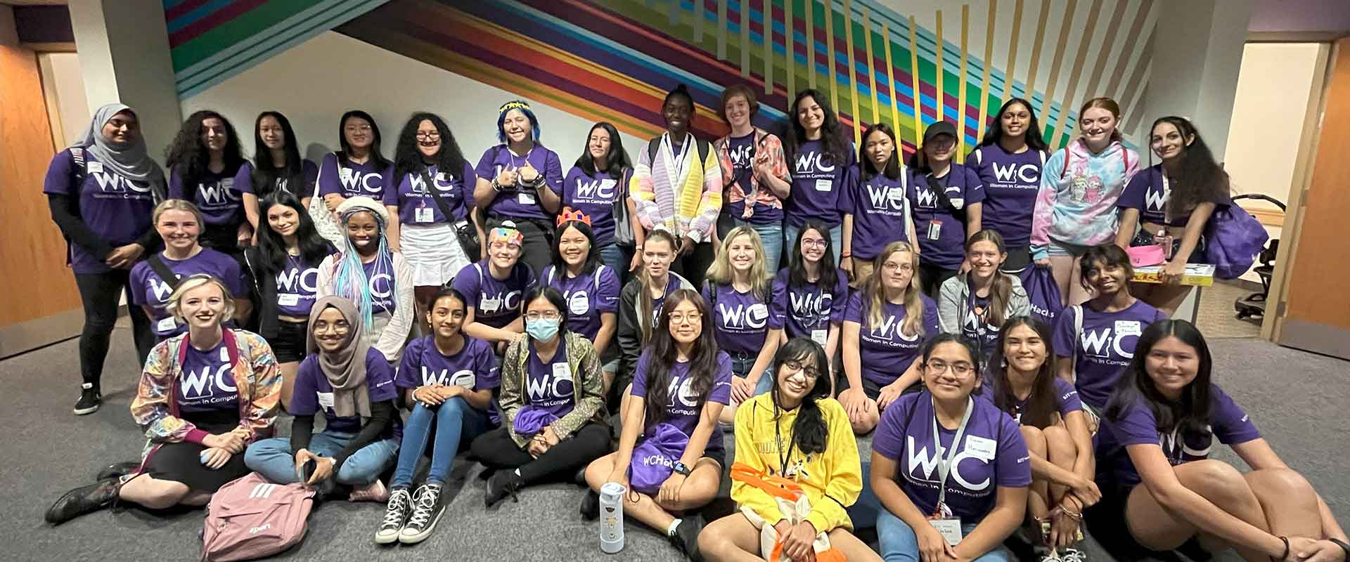 Students wearing purple shirts sit and pose together in front of a colorful wall mural.