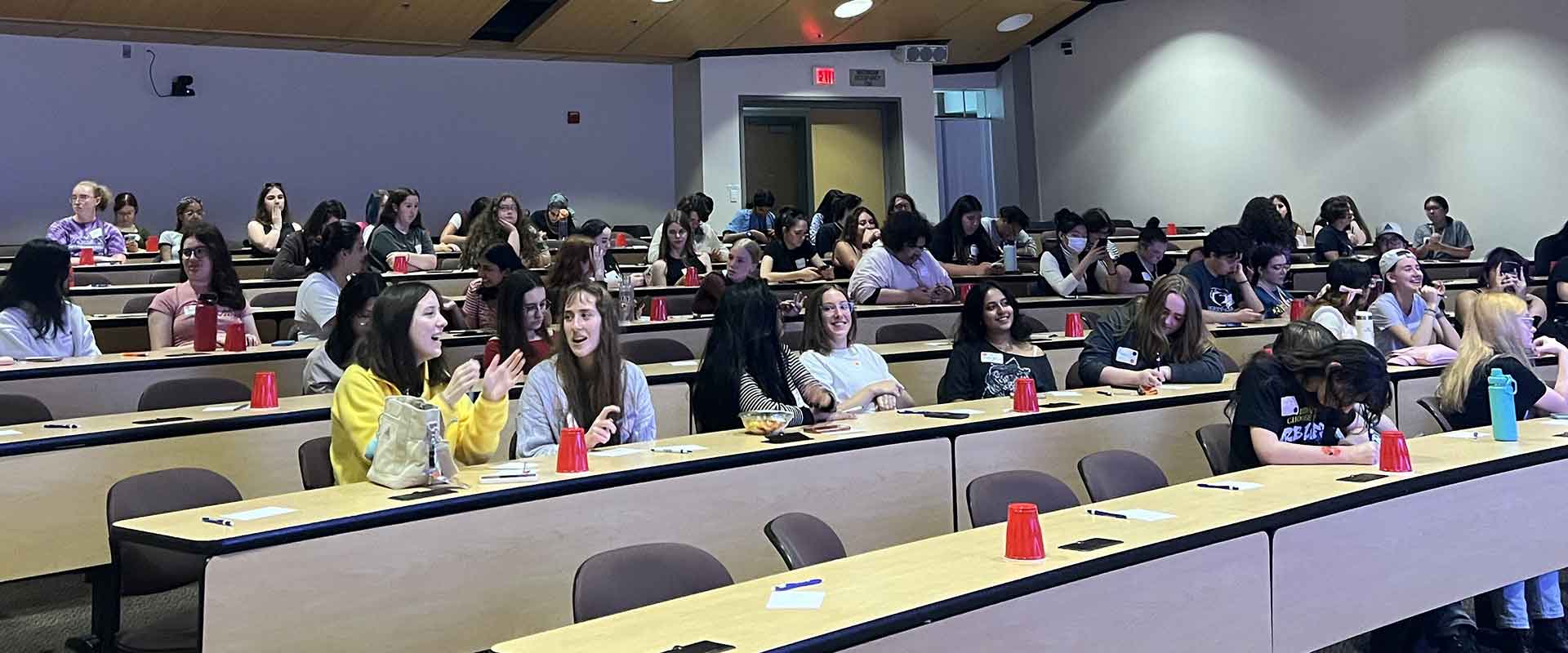 Students sit in tiered lecture hall seating while engaging in conversation and activities.