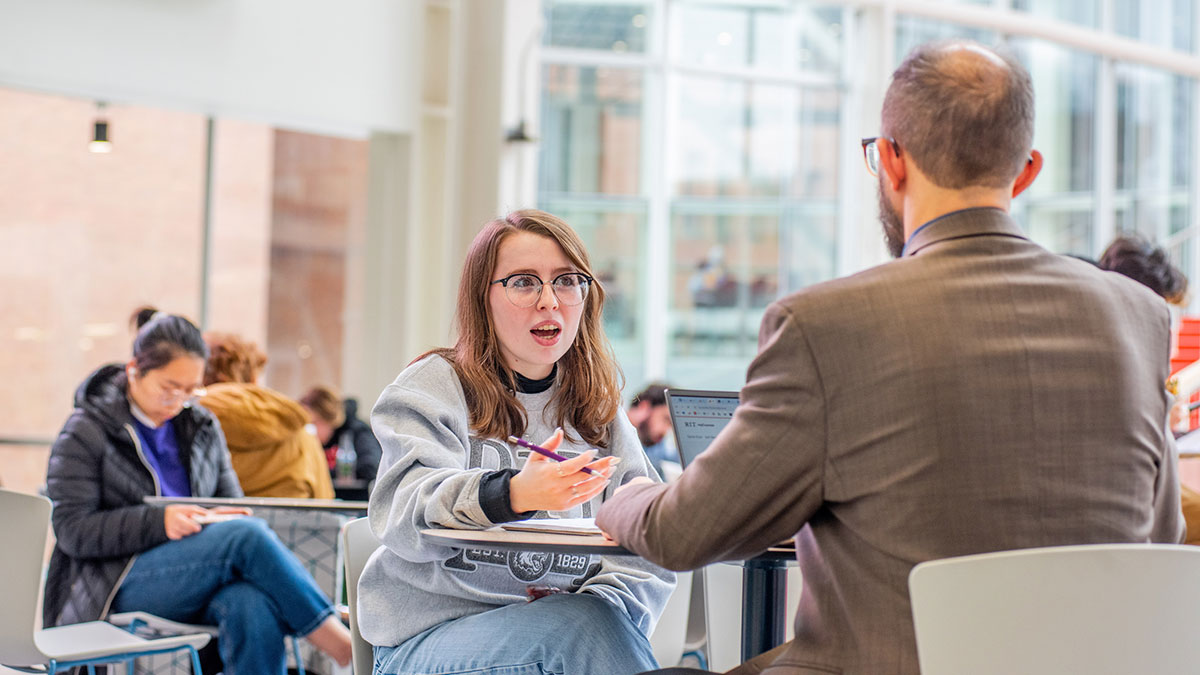 A student in an RIT sweatshirt gestures while speaking to a professional in a glass-walled campus building.