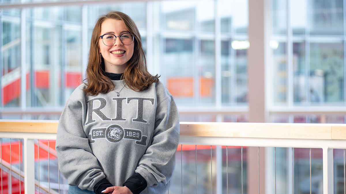 A smiling student wearing an RIT sweatshirt poses indoors in front of a glass-paneled balcony.