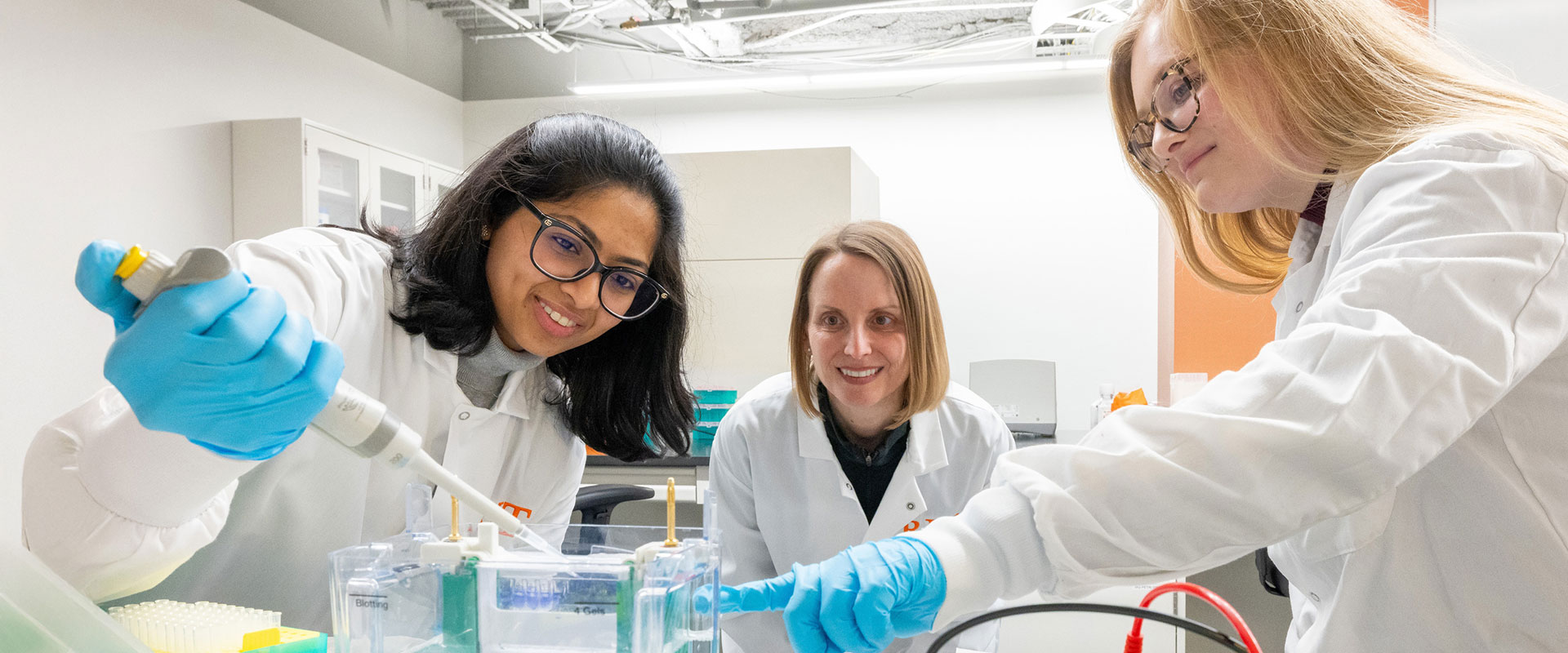 a researcher watches two students in a lab use a pipette.
