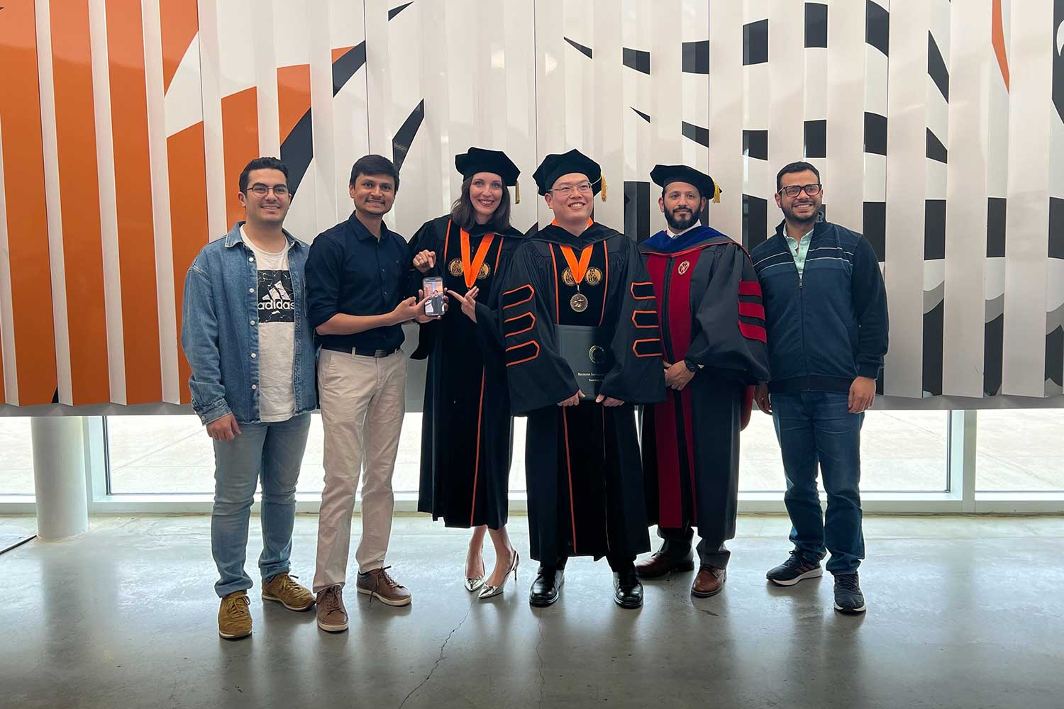 six people posing for a photo, with the three in the center dressed in graduation regalia.