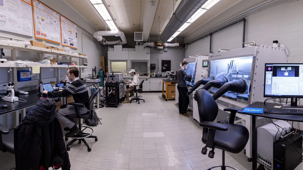 Wide view of a research laboratory with workbenches, microscopes, computers, overhead ventilation, and enclosed glovebox workstations. Three people are working at separate stations, including one seated at a lab bench, one seated near equipment at the back of the room, and one standing at a glovebox.