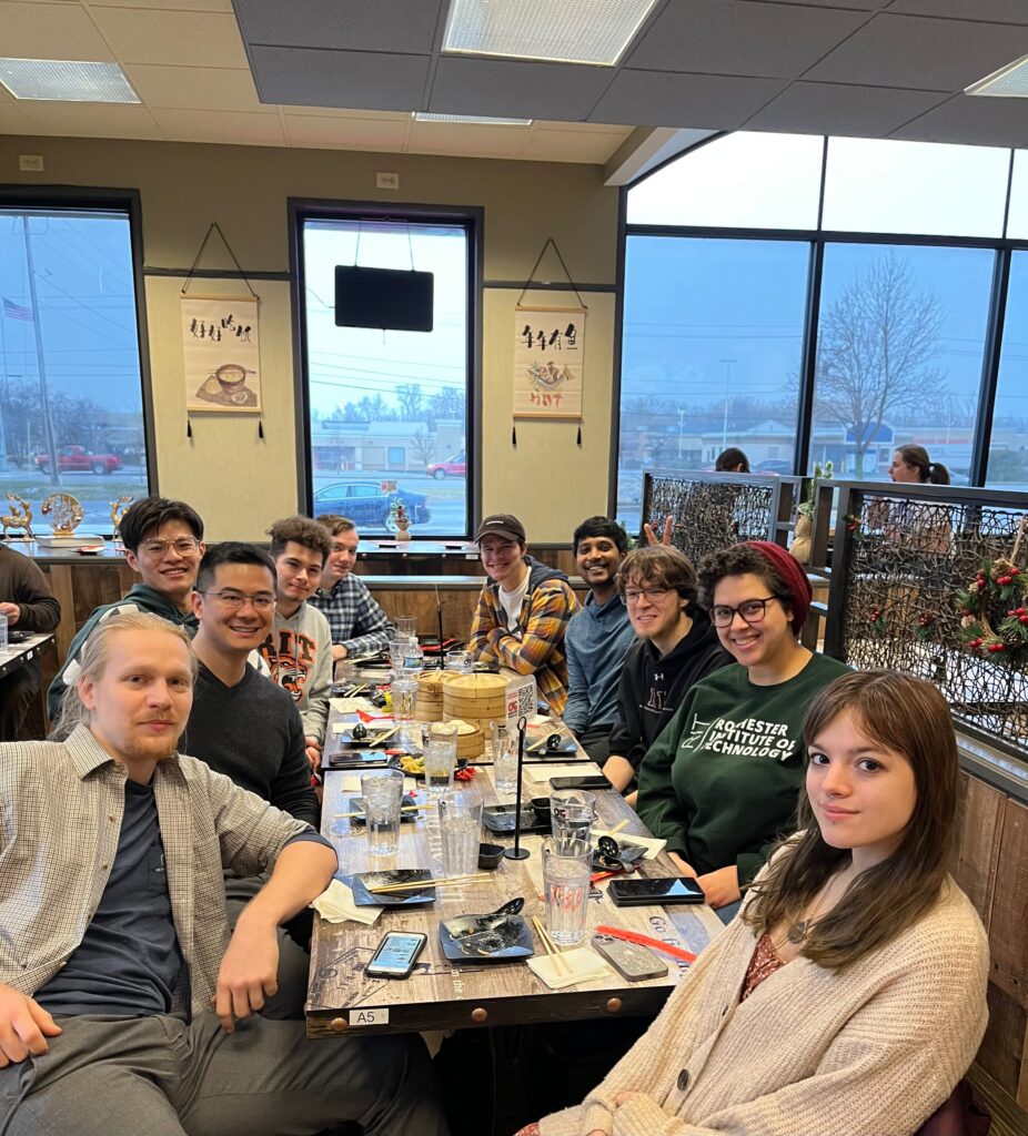 A group of people sit around a long restaurant table at OG Dumpling House with drinks, dishes, phones, and bamboo steamer baskets spread across the table. Large windows, wall hangings, holiday decorations, and other diners are visible in the background.