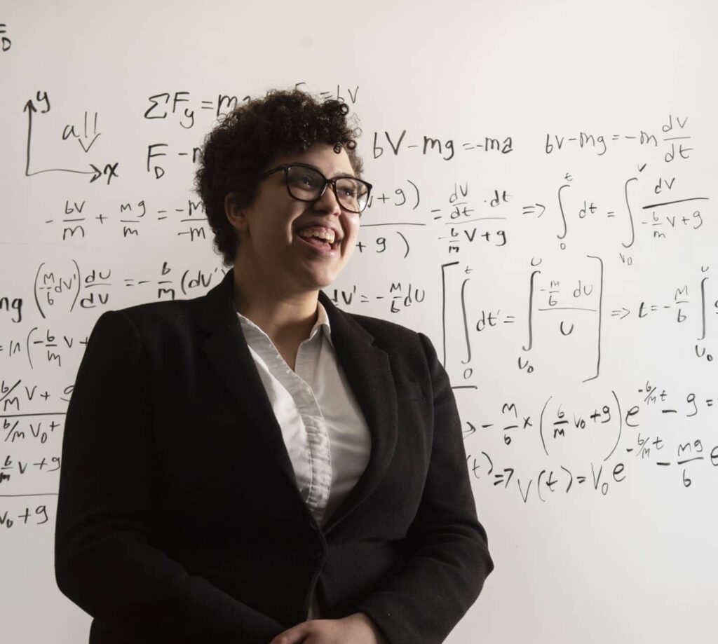 A person wearing glasses and a dark blazer stands in front of a whiteboard filled with handwritten physics equations and diagrams.
