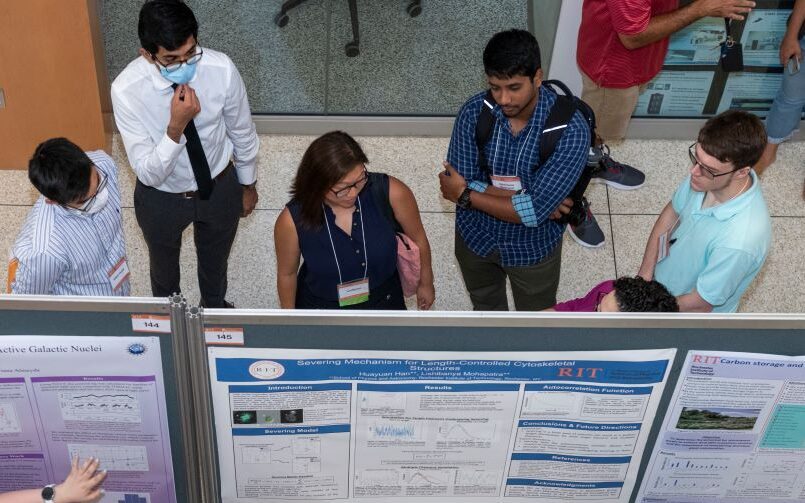 Overhead view of attendees gathered around research posters at the RIT Undergraduate Research Symposium. Several people stand in conversation near poster boards displaying project titles, graphs, and research summaries.