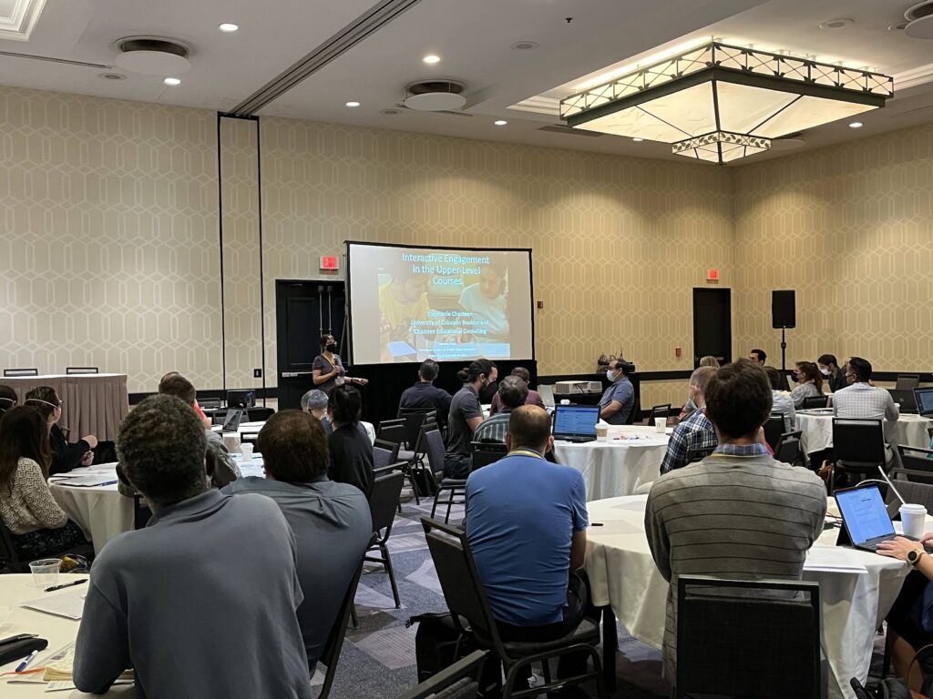 Attendees sit at round tables in a conference room during an AAPT Physics and Astronomy New Faculty Workshop presentation. A presenter stands near a projection screen showing a slide titled “Interactive Engagement in the Upper-level Courses,” while audience members use laptops and follow along.