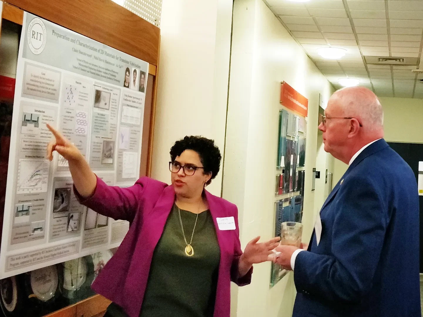 A presenter points to a research poster about preparation and characterization of 2D materials for transistor fabrication while speaking with an attendee in a hallway. The poster includes diagrams, graphs, and microscopy images related to 2D materials research.