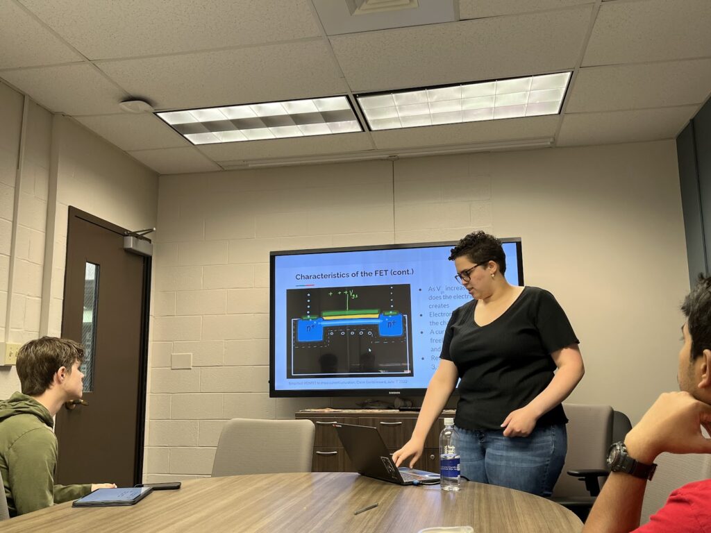 A person stands at the front of a conference room giving a presentation about field-effect transistor characteristics, with a hand-drawn transistor schematic displayed on a screen behind them. Two attendees sit at the table facing the presenter, with notebooks, a laptop, and a water bottle visible in the room.