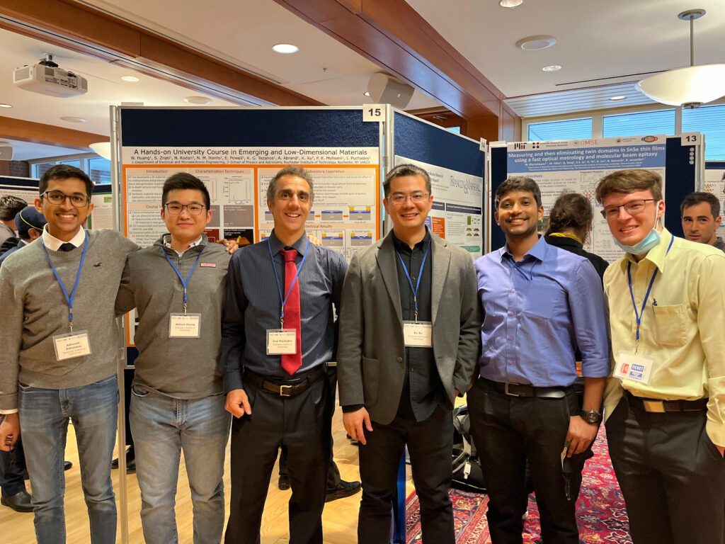 Six people wearing conference badges stand together in front of research poster displays at the 9th Graphene and Beyond Workshop. The indoor conference setting includes numbered poster boards, overhead lighting, and other attendees in the background.